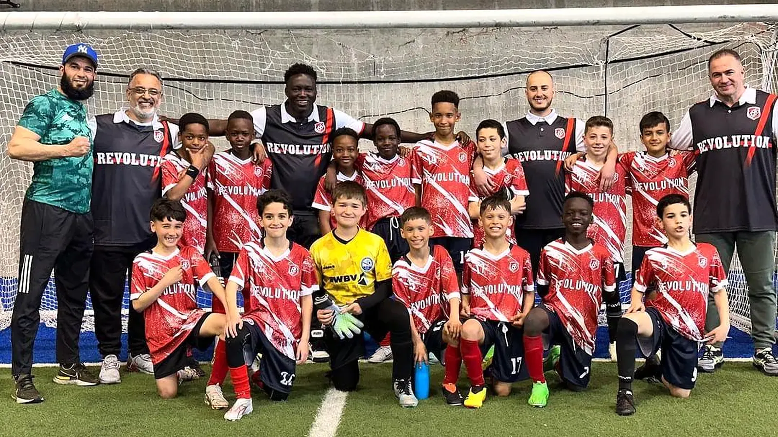 Équipe de jeunes joueurs Révolution réunie avec ses entraîneurs devant le but d'un terrain de soccer intérieur