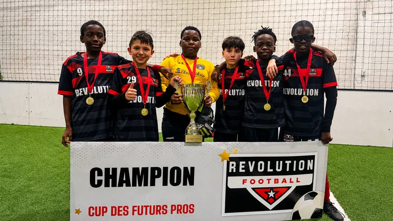 Joueurs de l'Académie Révolution en maillot noir et rouge et leur gardien en jaune, médailles d'or au cou, posant avec la coupe derrière la bannière Champion de la Cup des Futurs Pros, sur un terrain de soccer intérieur