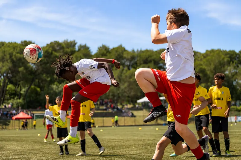 Joueur de l'Académie Révolution en maillot blanc et short rouge déviant le ballon de la tête lors d'un match de la Grinta Cup, un coéquipier en plein saut à ses côtés et les adversaires en jaune en arrière-plan